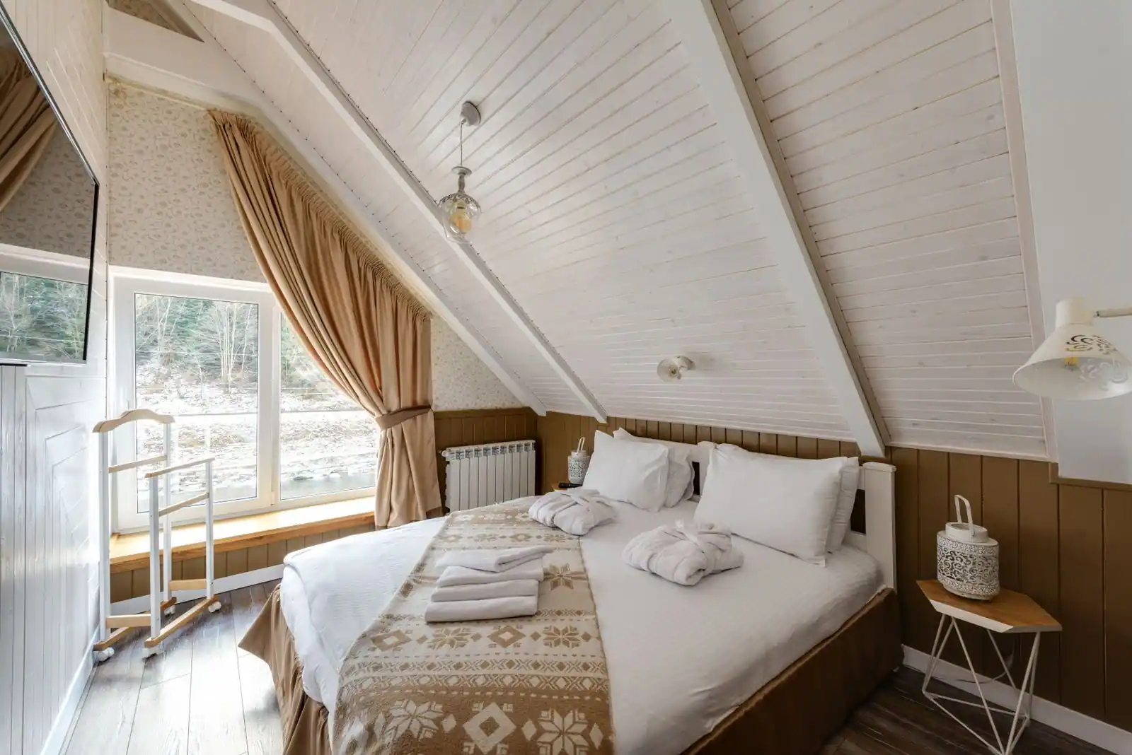Chambre mansardée au plafond en pente lambrissé de blanc, lit double installé sous le rampant et fenêtre habillée de rideaux
