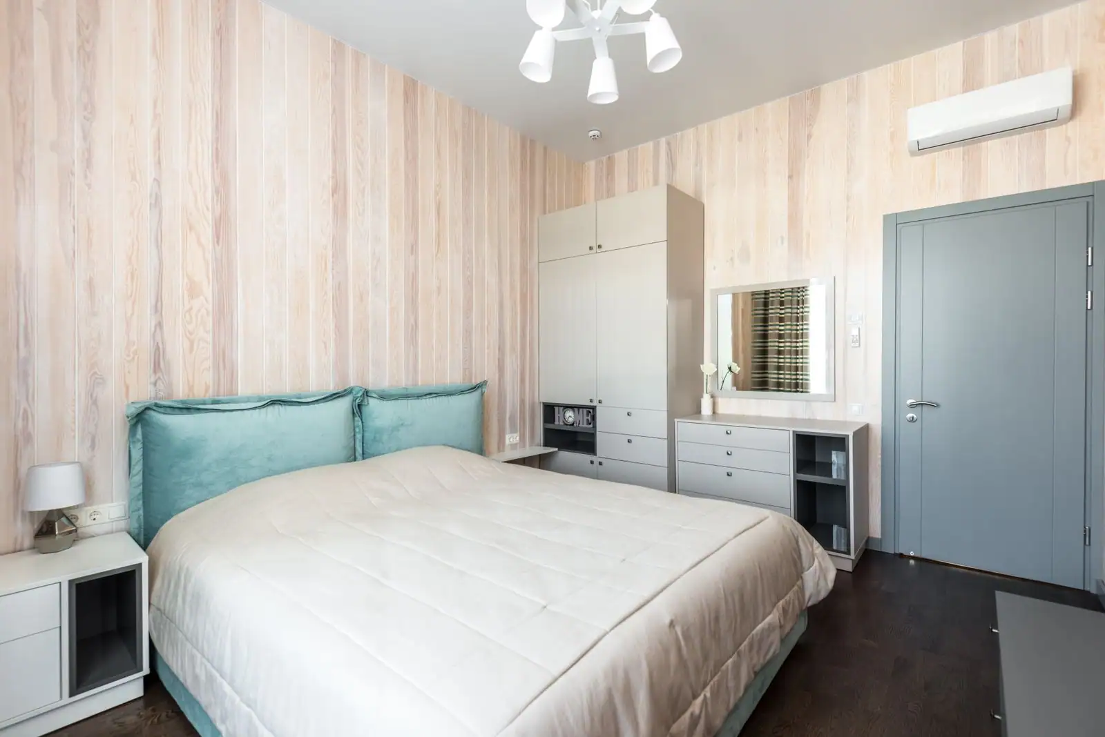 Chambre aménagée aux murs clairs en lames de bois, lit à tête capitonnée, armoire et miroir intégrés le long du mur