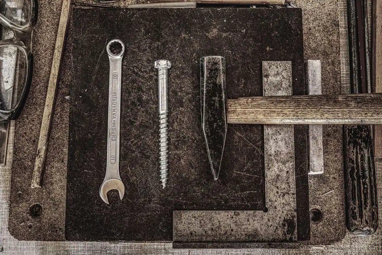 Outils de bricolage à plat — clé, grosse vis, burin et marteau — sur une surface d atelier patinée.