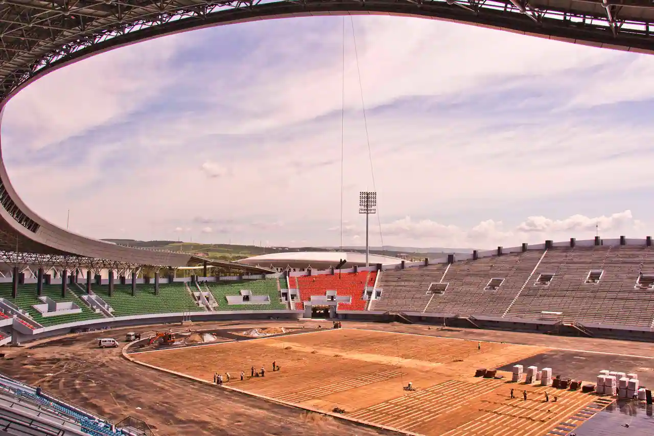 Grand stade en construction, gradins vides et pelouse en cours d'aménagement sous une vaste toiture courbe