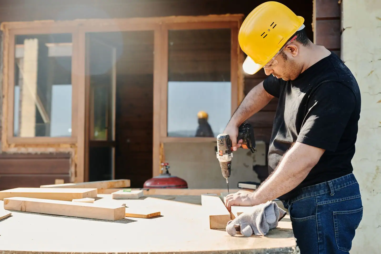 Un homme casque de chantier utilise une perceuse-visseuse sans fil sur une planche de bois lors de travaux de bricolage en extérieur.