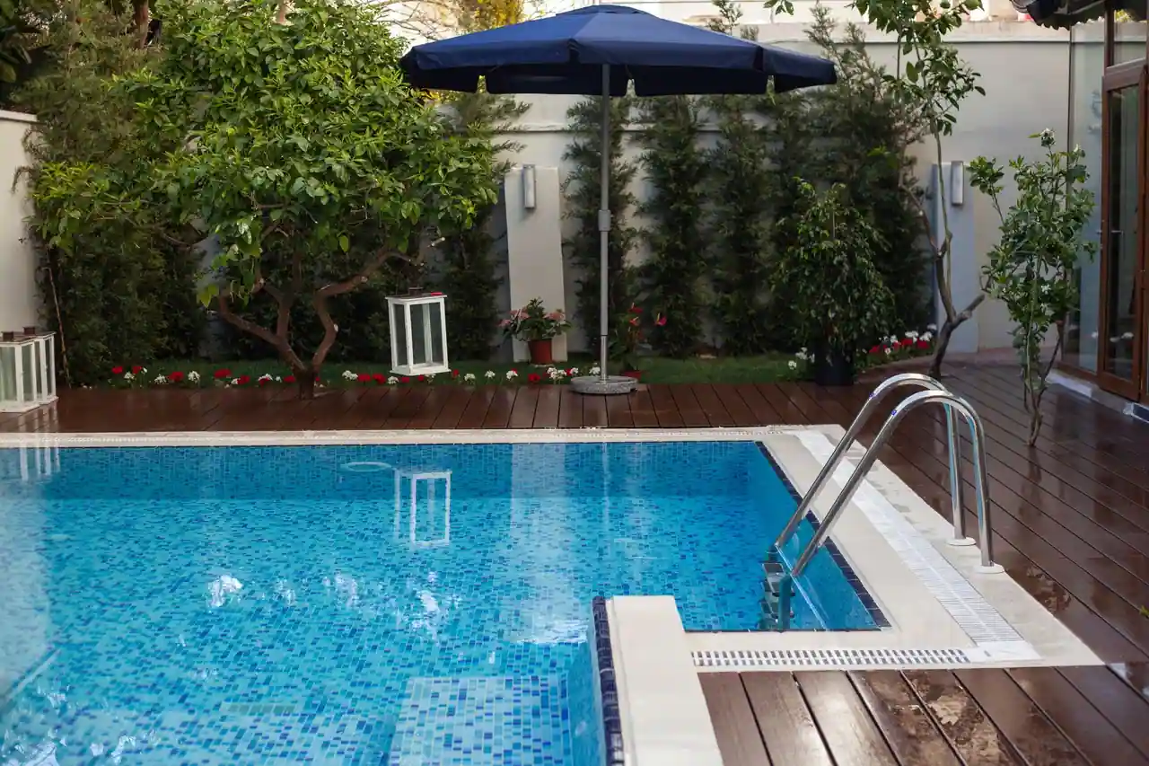 Petit bassin extérieur carrelé de bleu dans une cour close par des murs blancs, terrasse en bois et parasol.