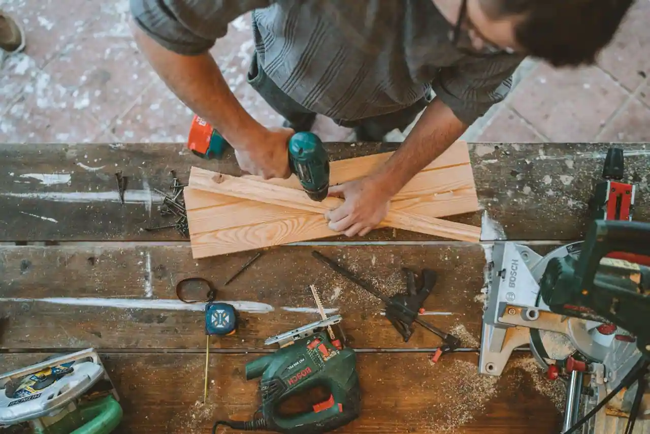 Mains perçant une planche de bois avec une perceuse sur un établi entouré d'outils de bricolage.